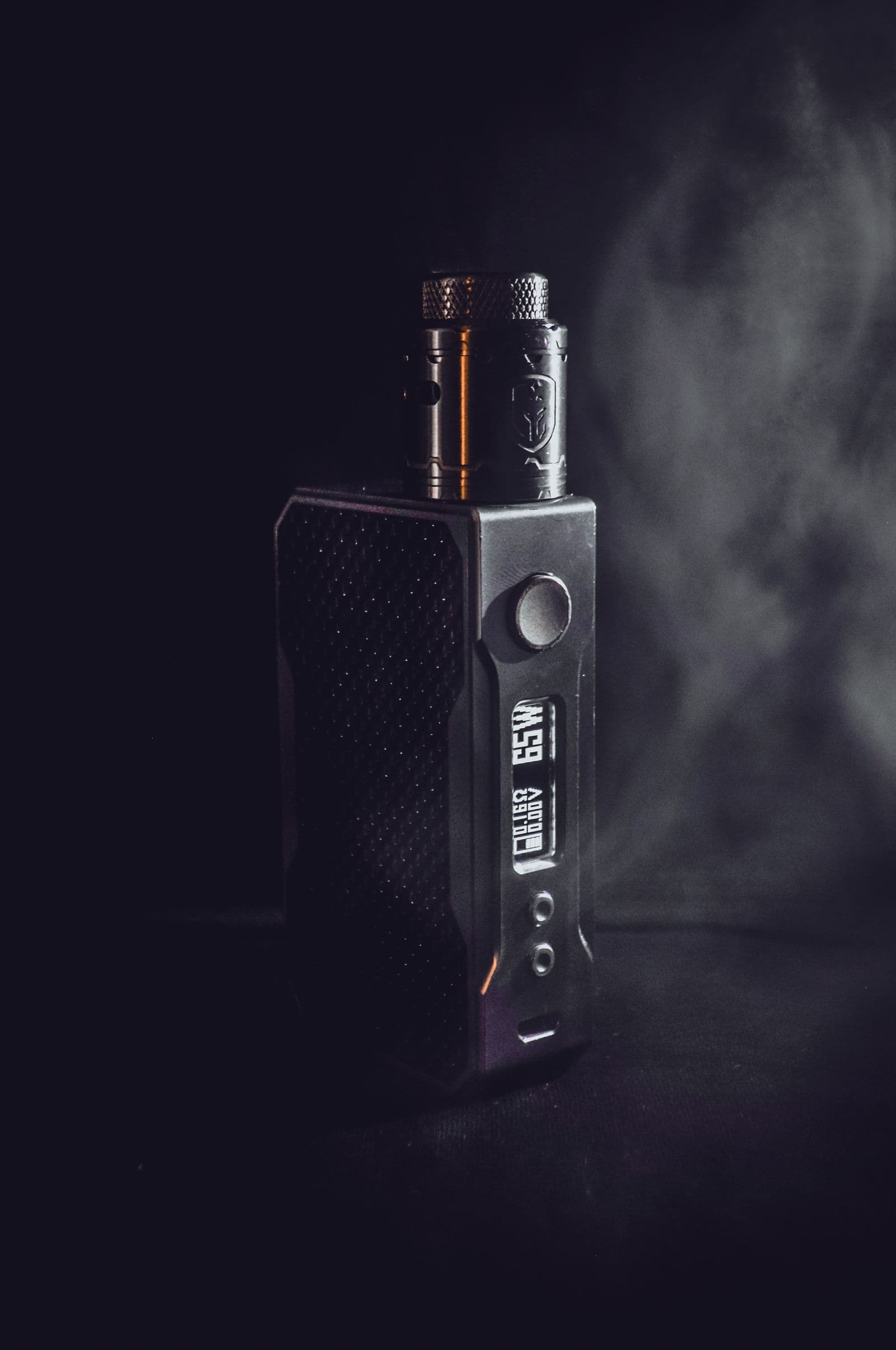 Box-mod vape device on a dark surface with a drift of vapor